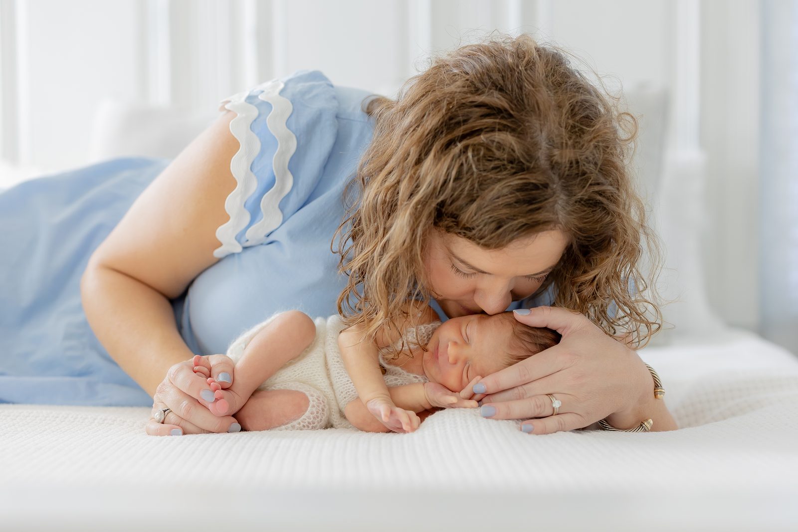 Mother in blue kissing her newborn's forehead on a white bed
