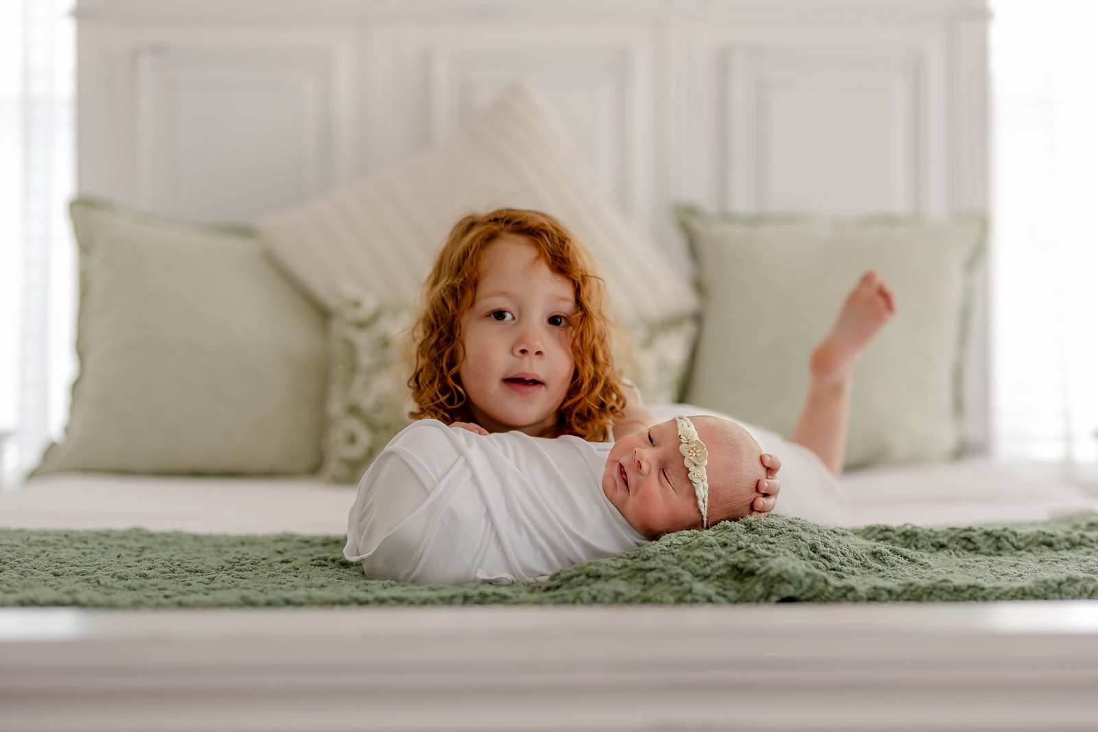 Big sister with red curls beside her swaddled newborn sibling in a floral headband