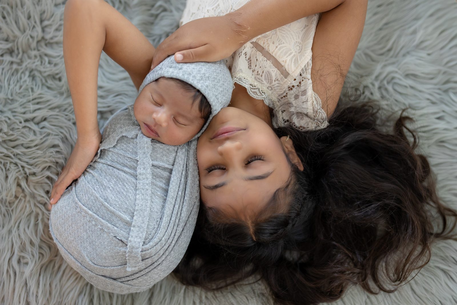 Mother and her swaddled newborn lying head to head on a soft grey rug