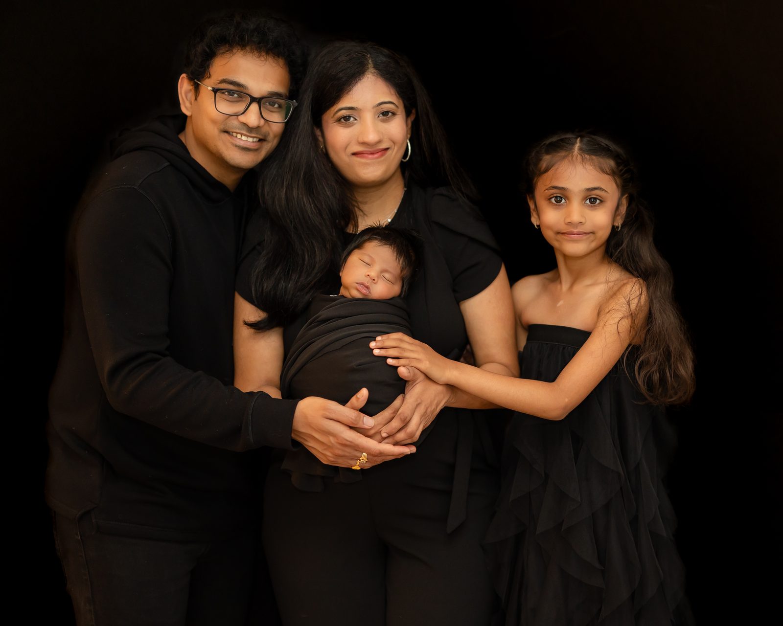 Family of four with their swaddled newborn on a black backdrop