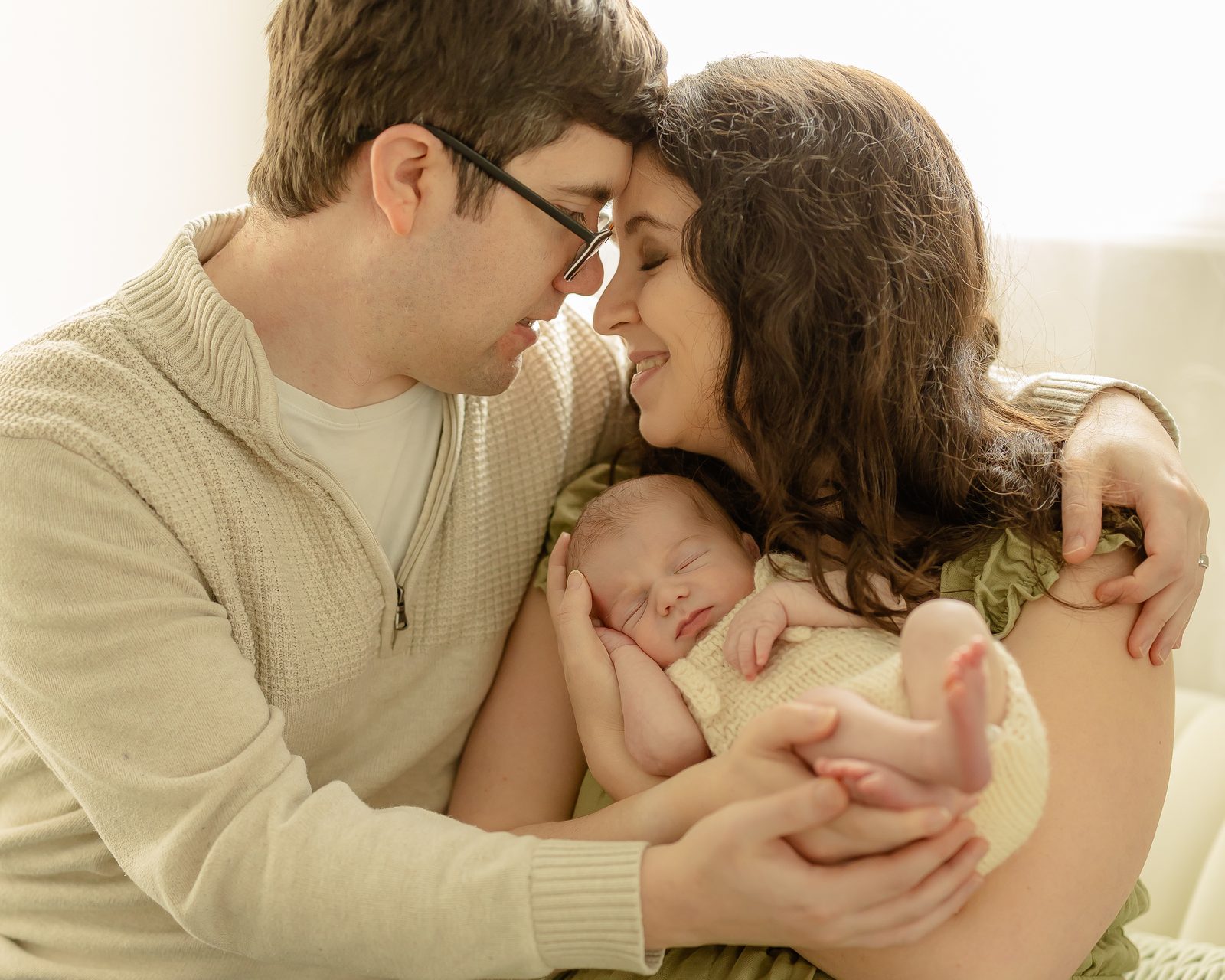 New parents foreheads together, cradling their newborn in cream knit